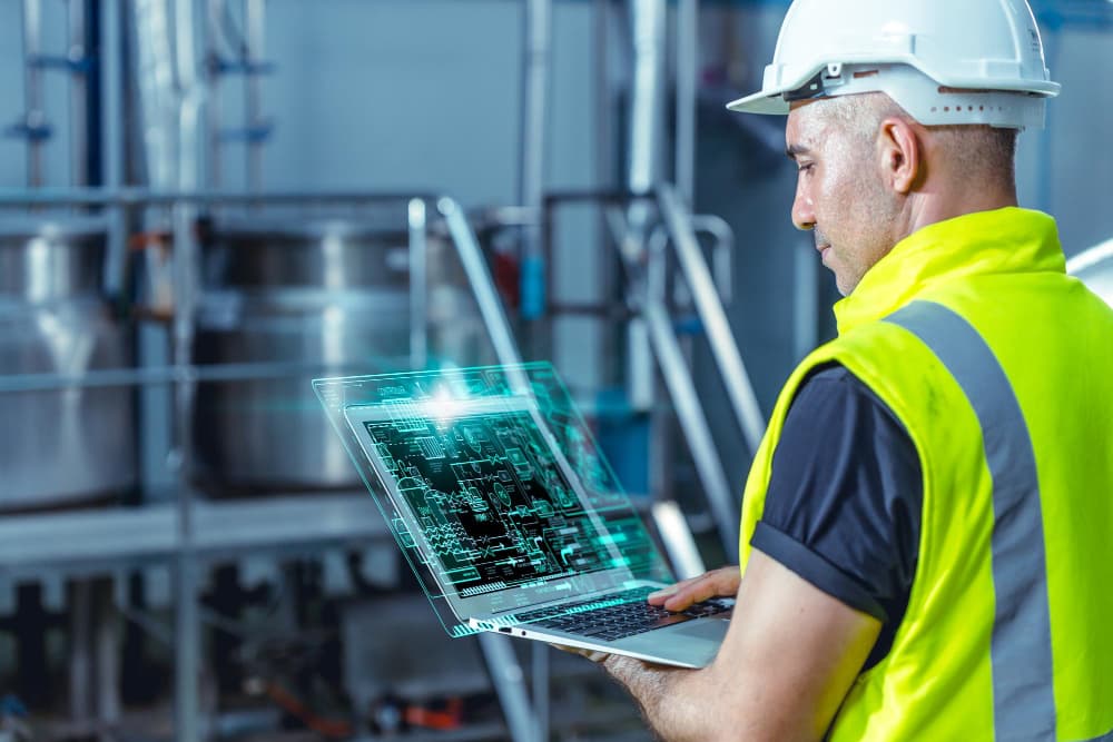 Mężczyzna w kamizelce odblaskowej i kasku ochronnym pracuje na laptopie z wyświetlonym schematem technicznym